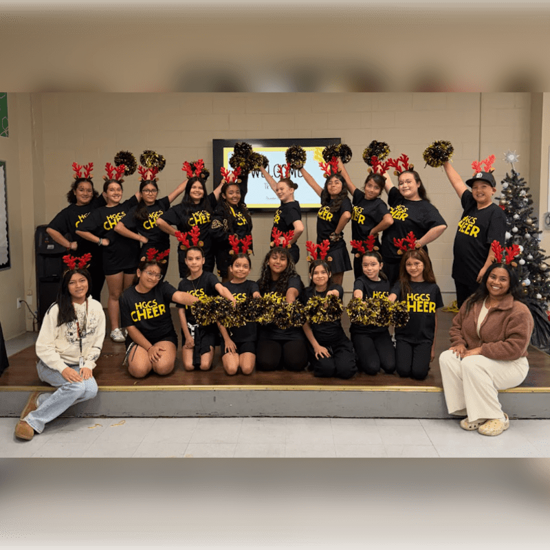 A group of young cheerleaders in black shirts and red reindeer antlers pose on a stage in front of a screen, with two individuals seated on the floor in the foreground.