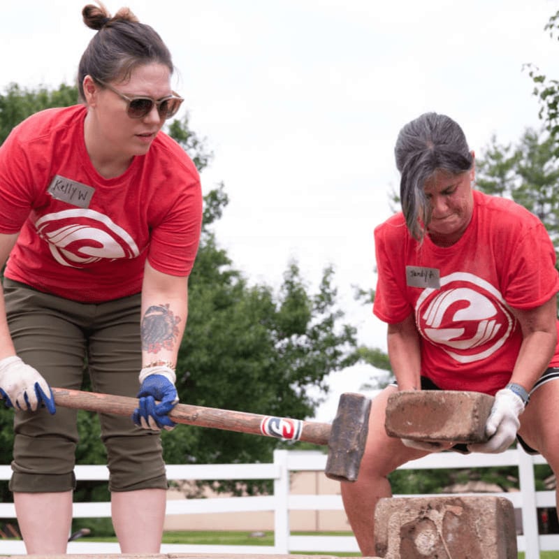 Two women in red shirts are smashing bricks with a sledgehammer in the foreground, with trees and a white fence in the background.
