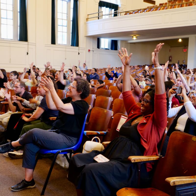 In the foreground, people are seated in rows of wooden chairs with their hands raised, while the background shows a large auditorium with more seating and a stage area.
