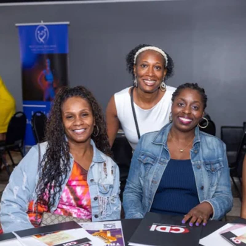 Three smiling women pose at a table with brochures in the foreground, while a blue banner hangs in the blurred background.