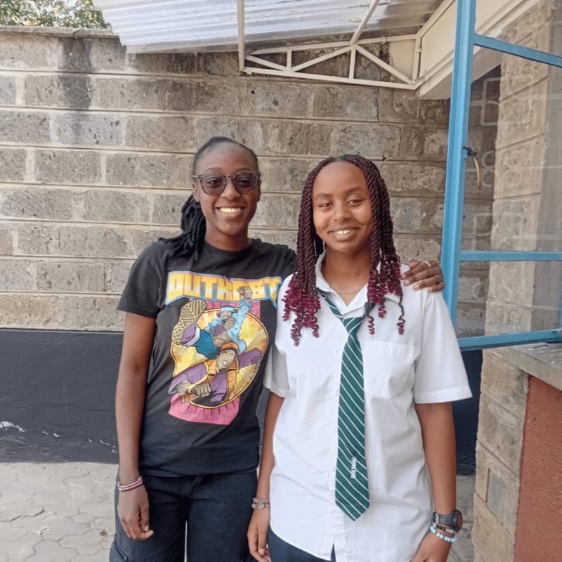 Two smiling young women stand in front of a brick wall, one in casual clothes and the other in a school uniform.