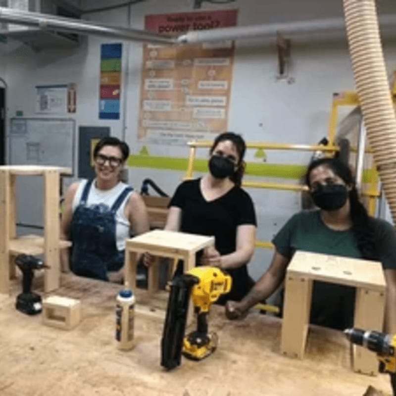 Three people in the foreground are working on small wooden stools with tools and glue, while the background shows a workshop with safety posters and equipment.