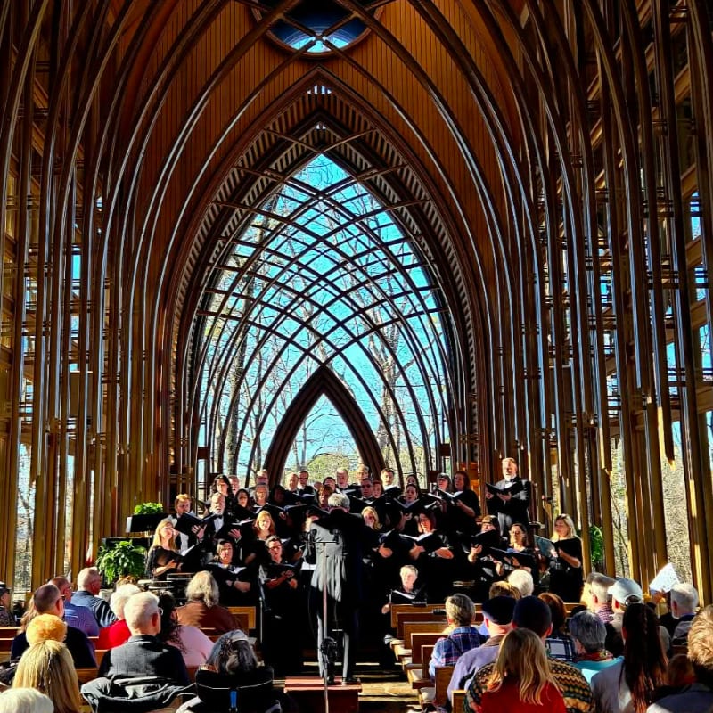 A choir performs in a sunlit chapel with a vaulted wooden ceiling, while an audience sits in pews in the foreground.