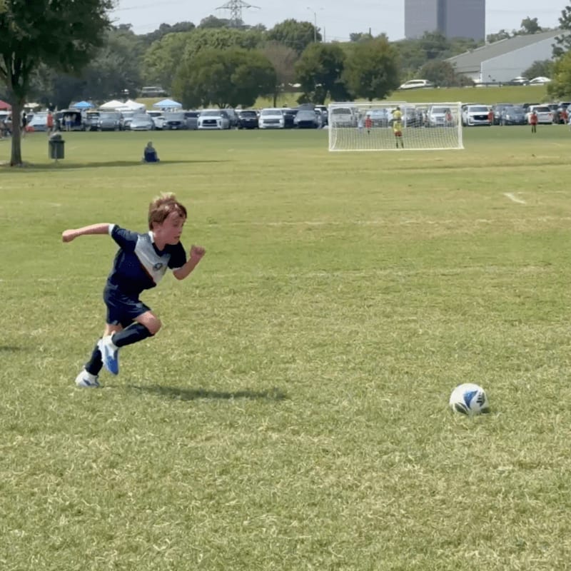 A young boy in a blue and white soccer uniform runs towards a soccer ball on a grassy field, with a soccer goal and parked cars visible in the background.