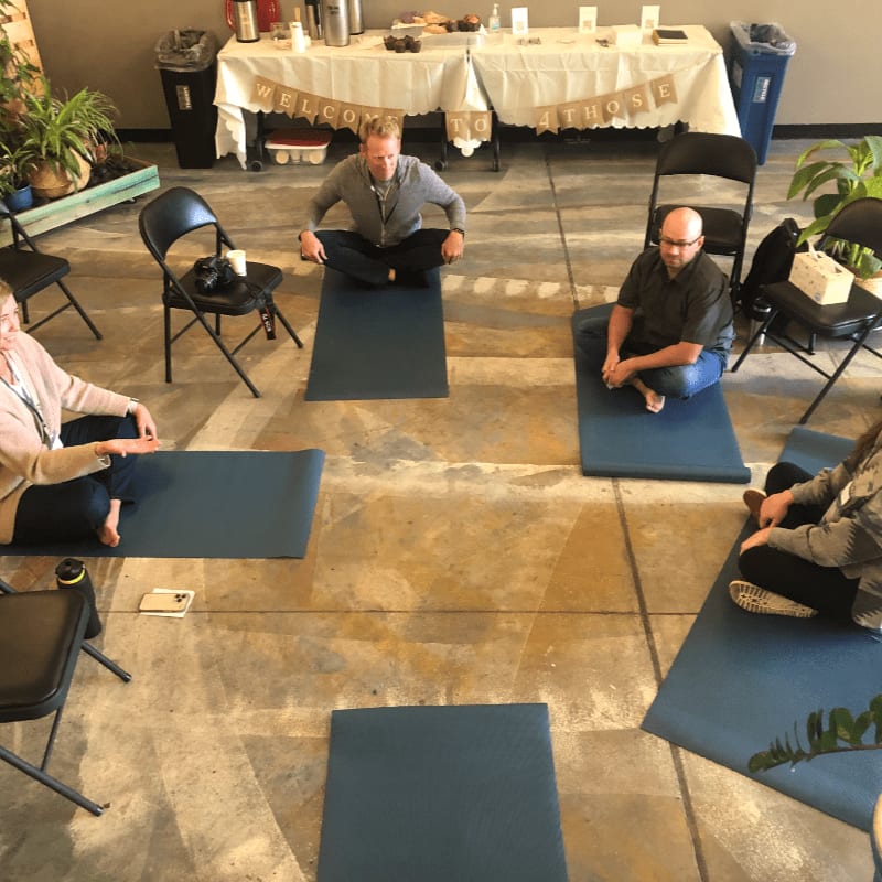 Four people sit in a circle on yoga mats in the foreground, while a table with a "Welcome" banner is visible in the background.