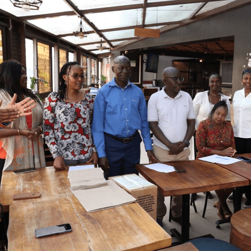 A group of people stand behind a table with papers and phones in the foreground, while a restaurant interior with windows and lighting is visible in the background.