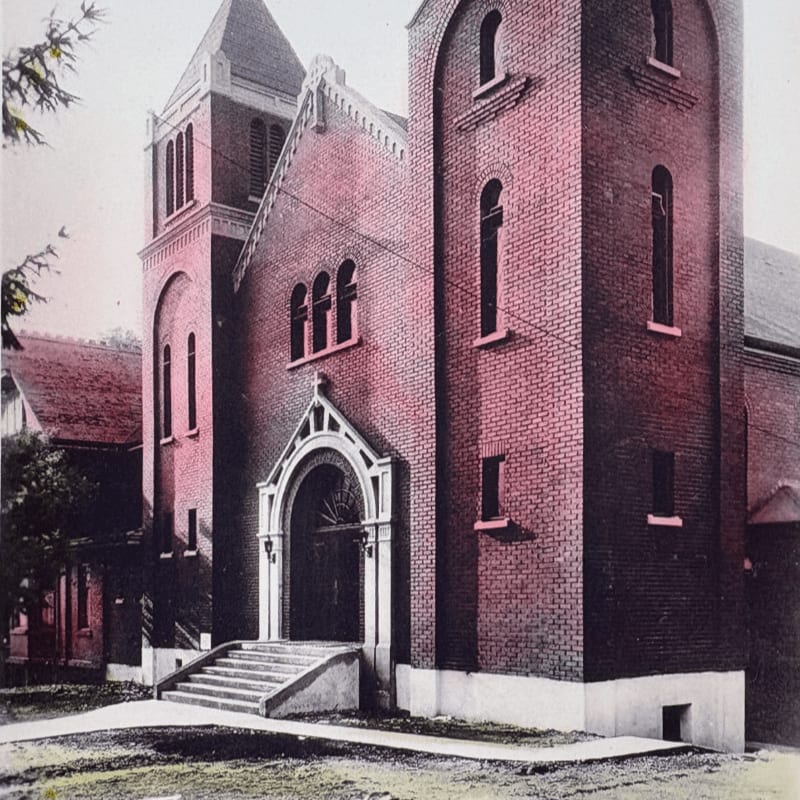 A brick church with a prominent entrance and bell tower stands against a backdrop of trees and other buildings.