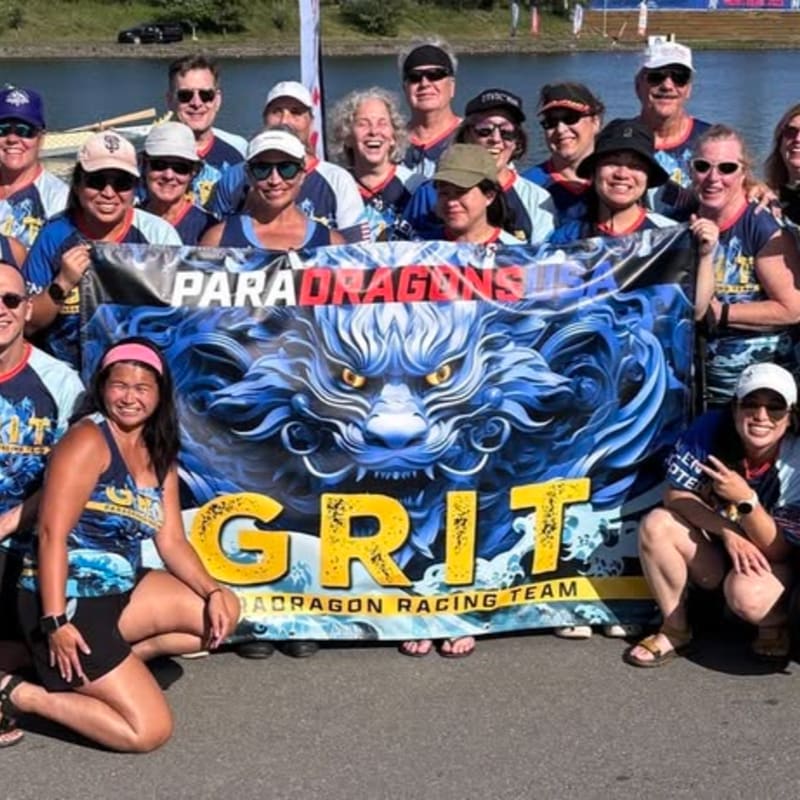 A group of smiling people in matching shirts pose in front of a banner featuring a dragon and the word "GRIT" with a body of water and trees in the background.