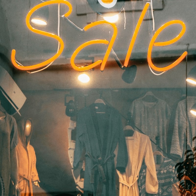 An orange neon "Sale" sign glows above a window display of clothing, including robes and sweaters, with a glimpse of an air conditioner unit in the background.