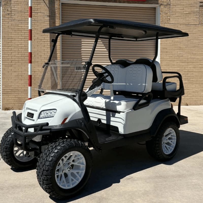 A white four-seater golf cart with black accents sits on a concrete surface in front of a brick building.