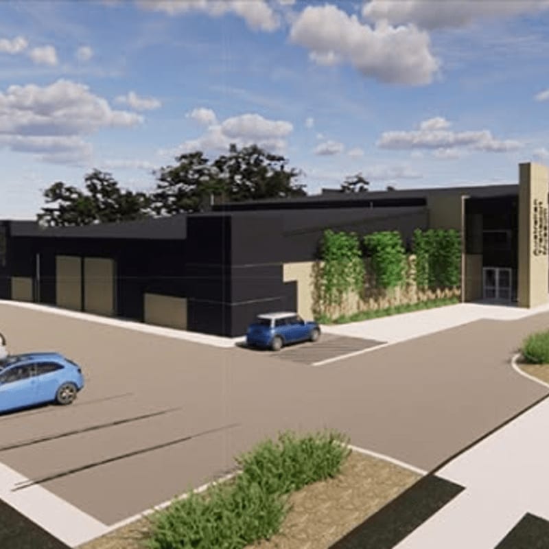 Two blue cars are parked in a lot in front of a modern building with black and tan facades, lush greenery, and a bright blue sky with scattered clouds.