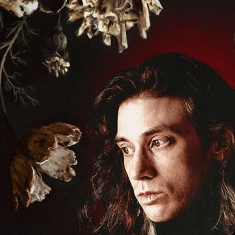 A man with long dark hair gazes to the side against a dark background with dried flowers in the foreground.