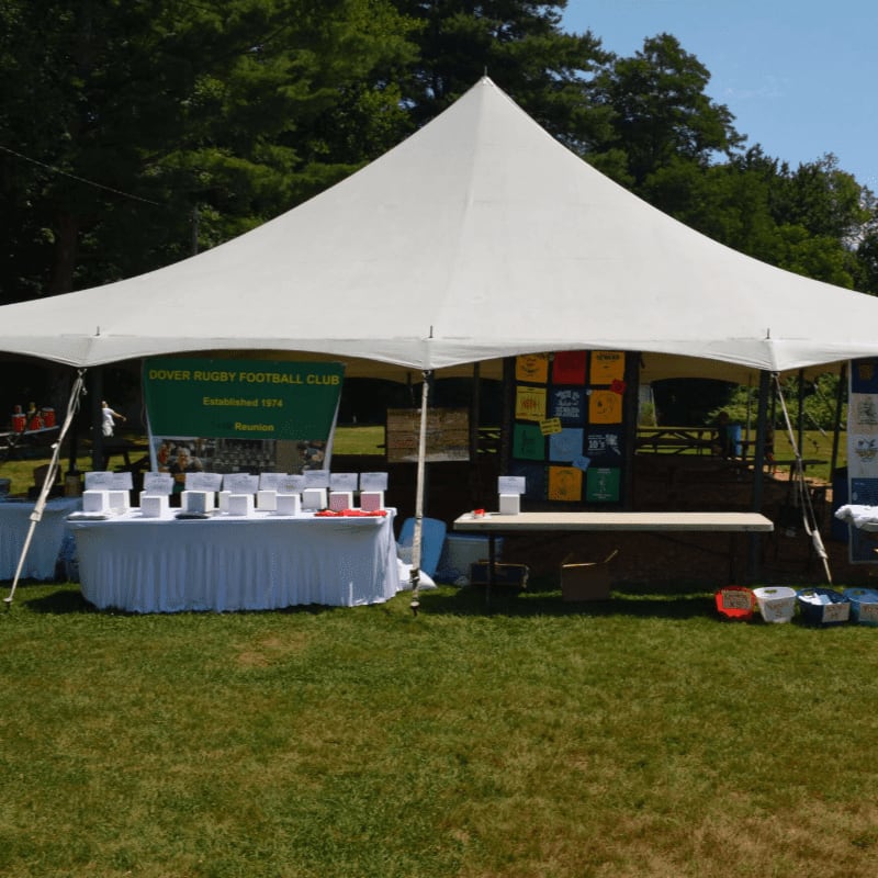 A white tent stands on a grassy field, displaying tables of items and colorful t-shirts, with trees and a clear sky in the background.