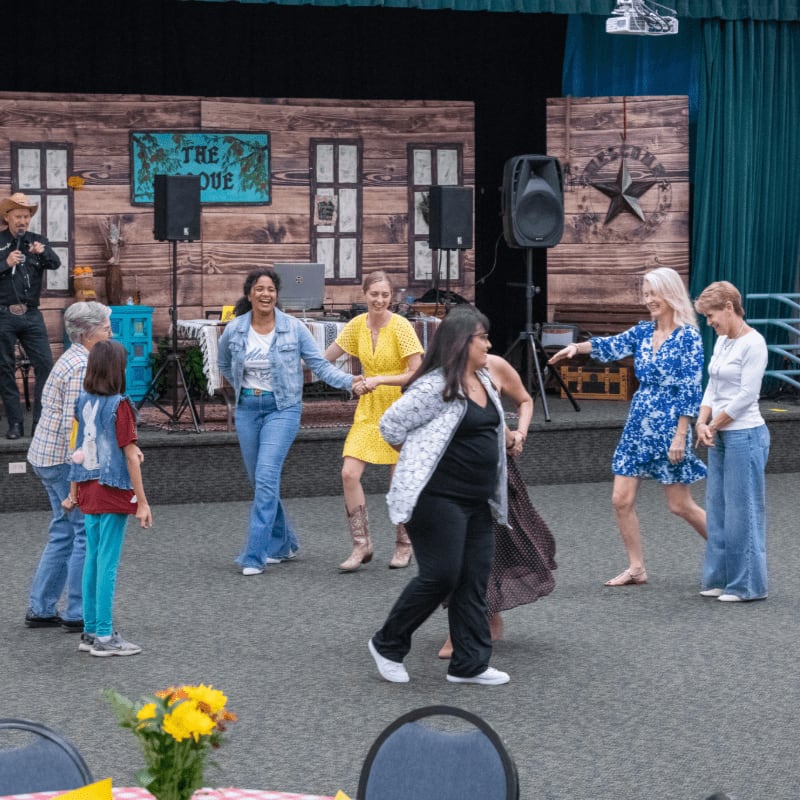 A group of people in casual attire dance on a gray floor in front of a rustic wooden stage backdrop.