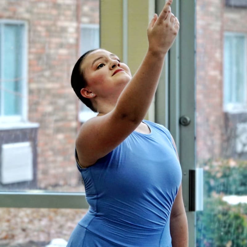 A dancer in a blue leotard reaches upwards with her right arm, with a blurred background of windows and brick buildings.