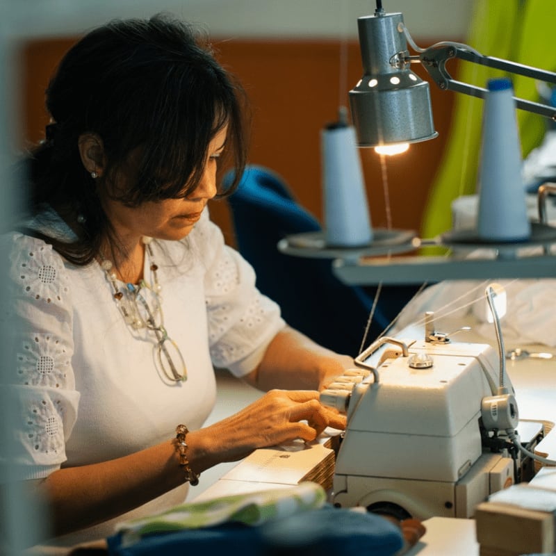 A woman in a white shirt sews at a machine, with spools of thread and fabric visible in the background.