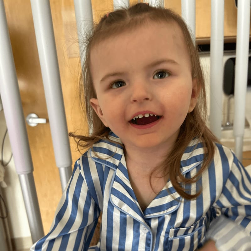 A young child in striped pajamas smiles in the foreground, with a hospital bed railing and a door visible in the background.