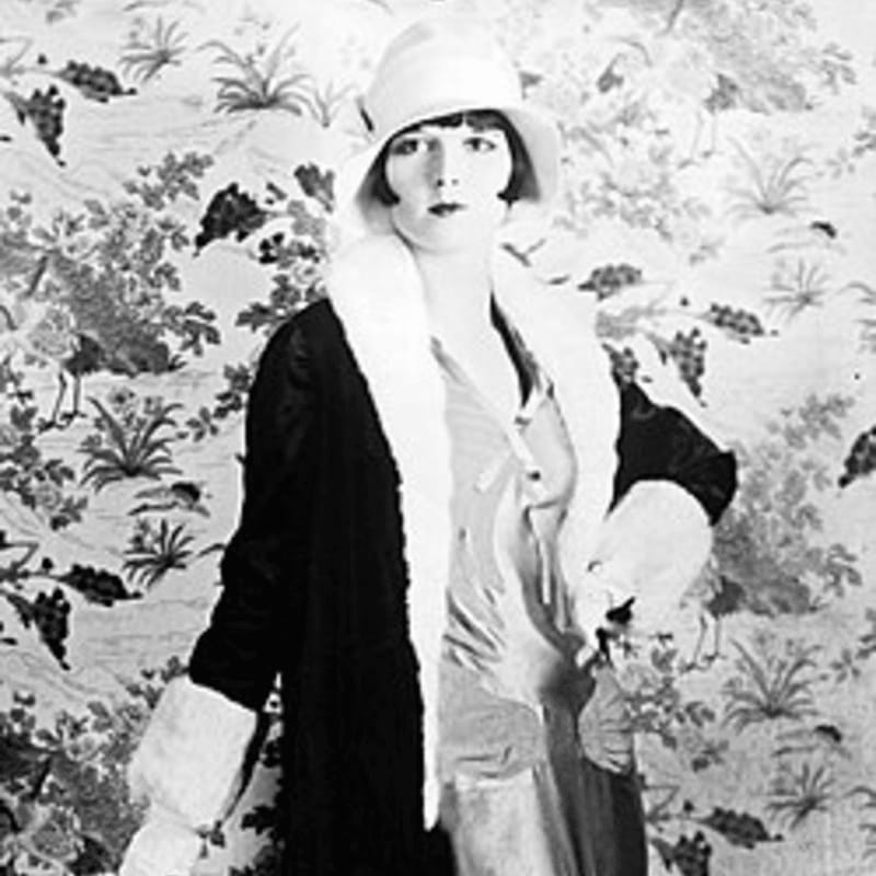 A woman in a cloche hat and fur-trimmed coat stands against a patterned wallpaper.