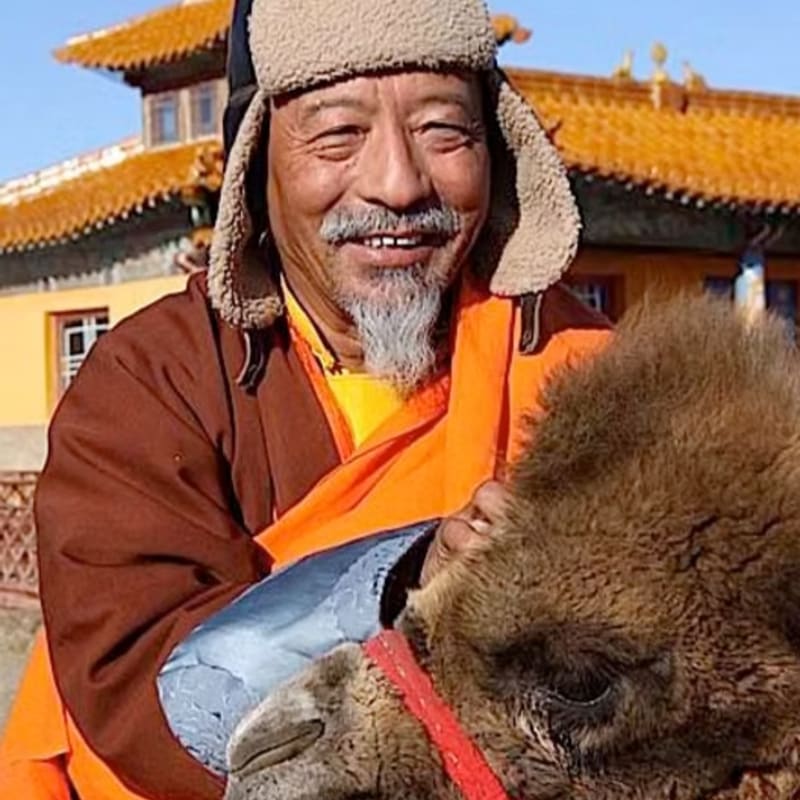 A smiling man in traditional robes and a fur hat poses with a camel in front of a yellow building with an orange roof.