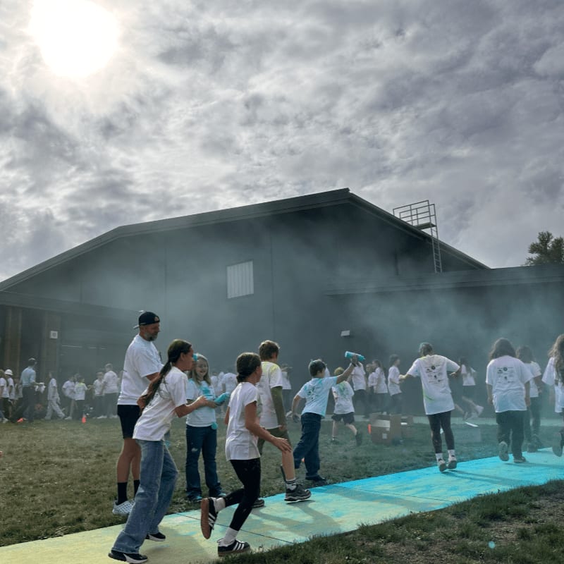 Children in white shirts run through blue and green powder in front of a large building under a cloudy sky.