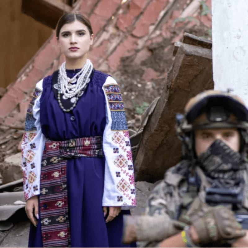 A woman in traditional Ukrainian attire stands in the foreground against a backdrop of a destroyed building, with a blurred soldier in military gear to her right.