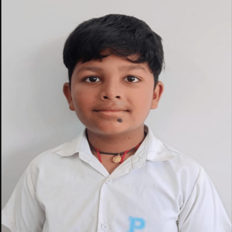 A young boy with dark hair and a mole on his cheek, wearing a white collared shirt, stands against a plain white background.