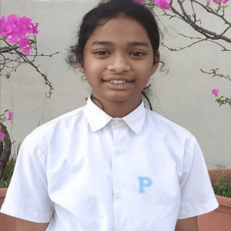 A young girl in a white collared shirt stands in front of a wall with pink flowers and branches.