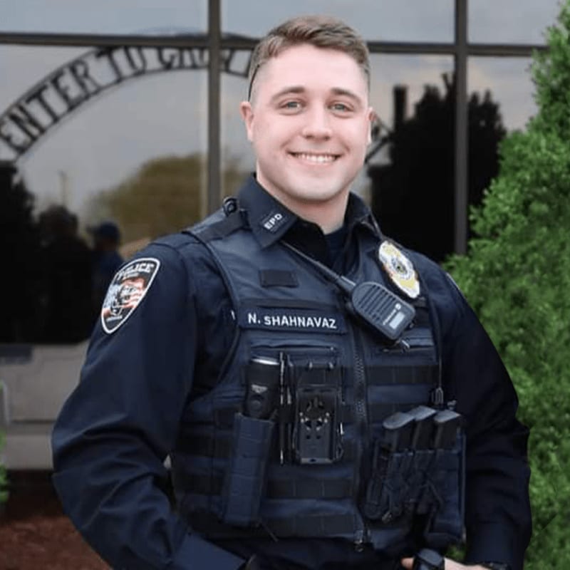 A smiling police officer in uniform stands in front of a building with an arched entrance.