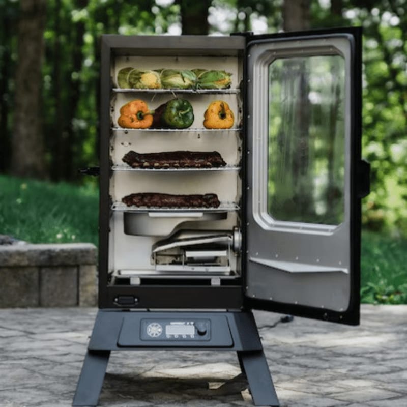 A smoker filled with ribs, corn, and peppers stands on a patio with a blurred green, wooded background.