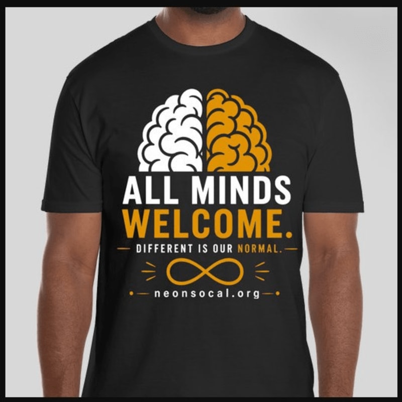A black t-shirt with a graphic of a split brain and text reads "ALL MINDS WELCOME. DIFFERENT IS OUR NORMAL." against a plain gray background.
