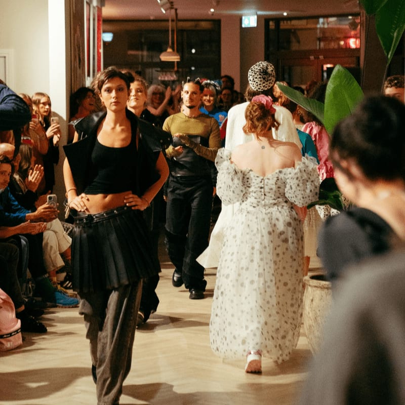 Models walk down a runway in fashionable outfits while an audience watches in the background.