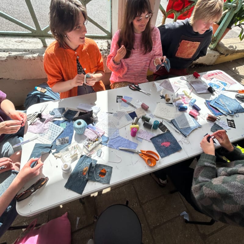In the foreground, a group of people are gathered around a table covered in fabric scraps, needles, and thread, while in the background, a sunny outdoor setting with a railing and plants is visible.