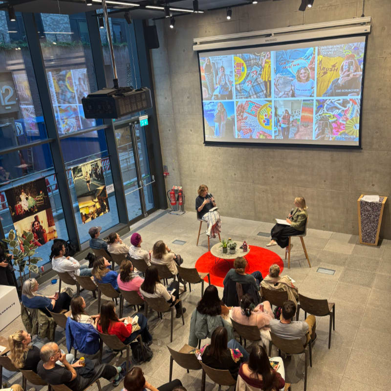 In the foreground, an audience sits facing two women on a stage, while the background features a large screen displaying a collage of images and a modern, glass-fronted building.
