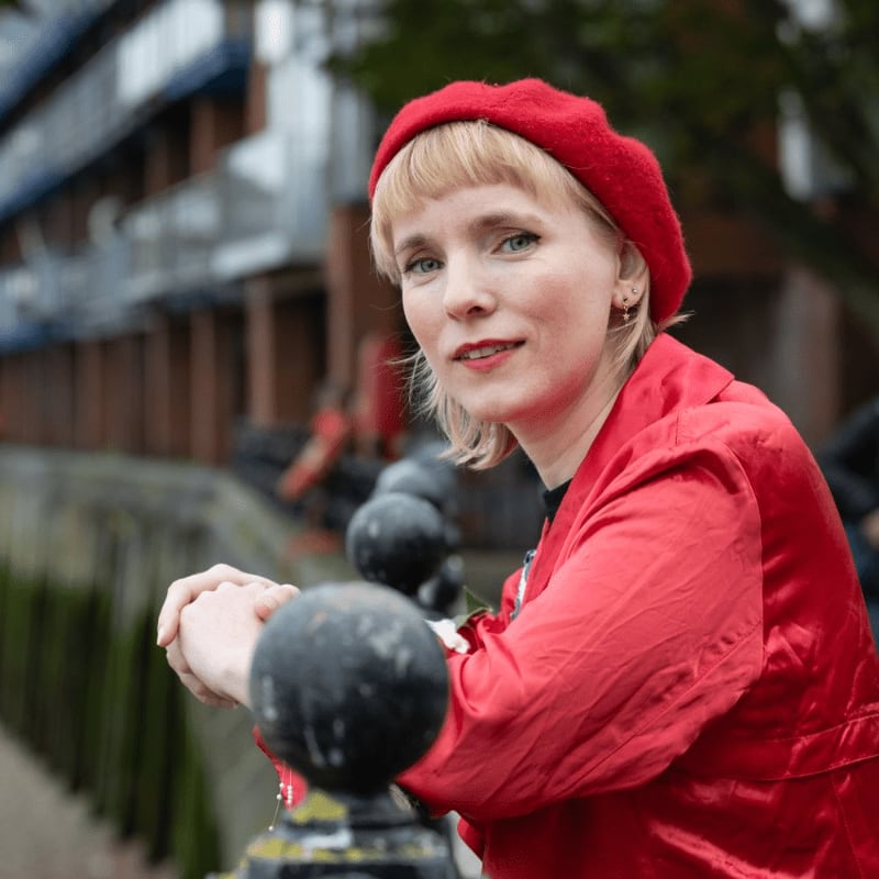 A woman in a red beret and jacket leans on a railing in the foreground, with a blurred urban background of buildings and trees.