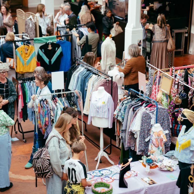 People browse racks of colorful clothing and accessories at an indoor market, with more shoppers and stalls visible in the background.