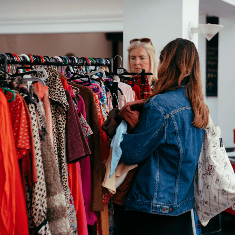 A woman in a denim jacket browses a rack of colorful clothes in the foreground, while another woman looks on in the blurred background.