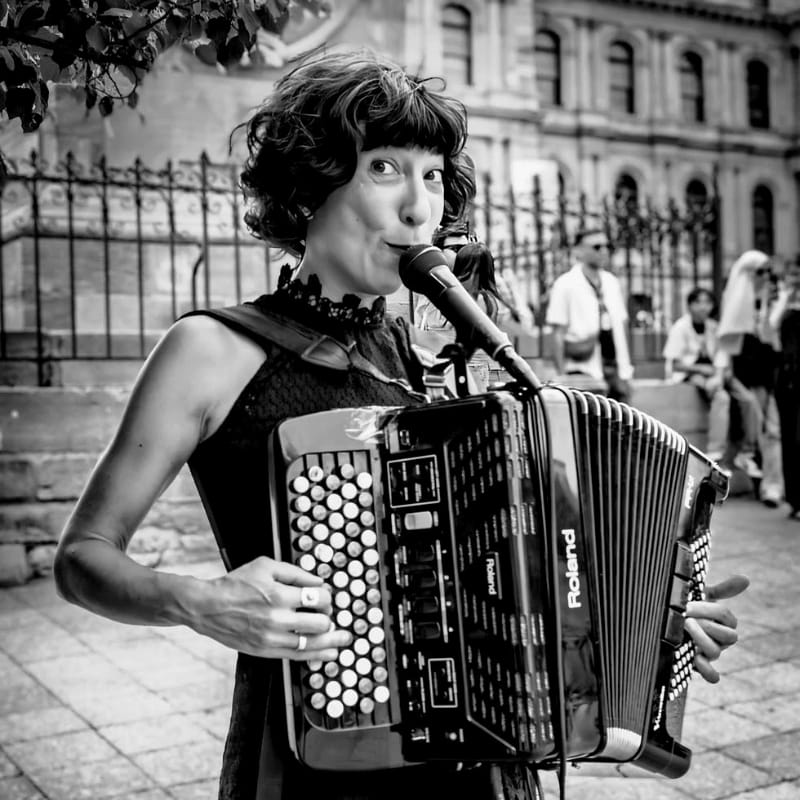 A woman plays an accordion in the foreground, with a blurred background of people and a building.