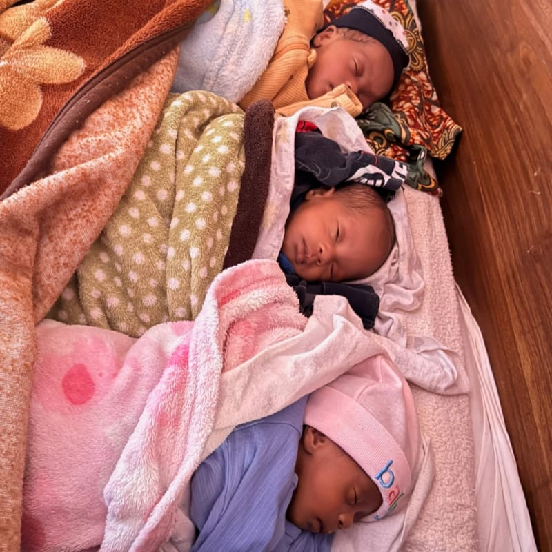 Three newborns sleep soundly, nestled amongst colorful blankets and hats, in a wooden crib.