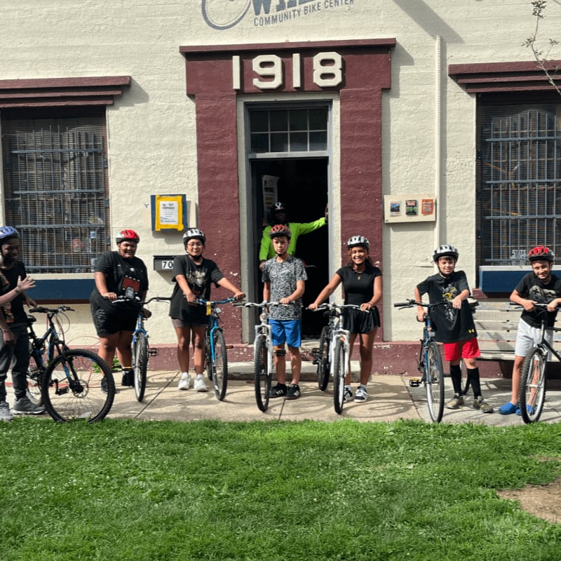 A group of children with bicycles stand in front of a building with "1918" above the entrance, while a grassy lawn is in the foreground.