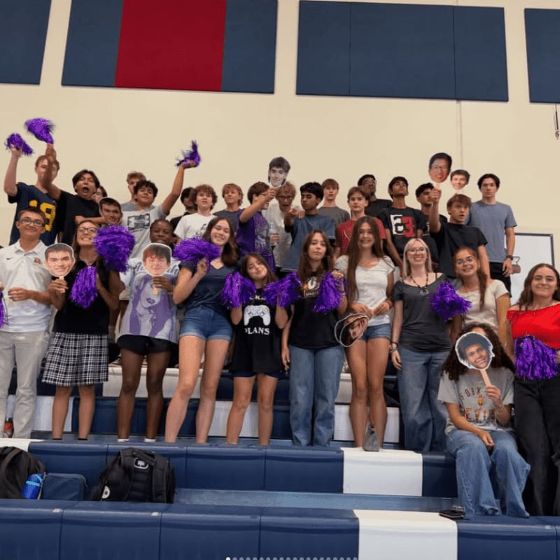 A group of teenagers in the foreground hold up cutout faces and purple pom-poms, while in the background, more students stand on bleachers in a gymnasium.