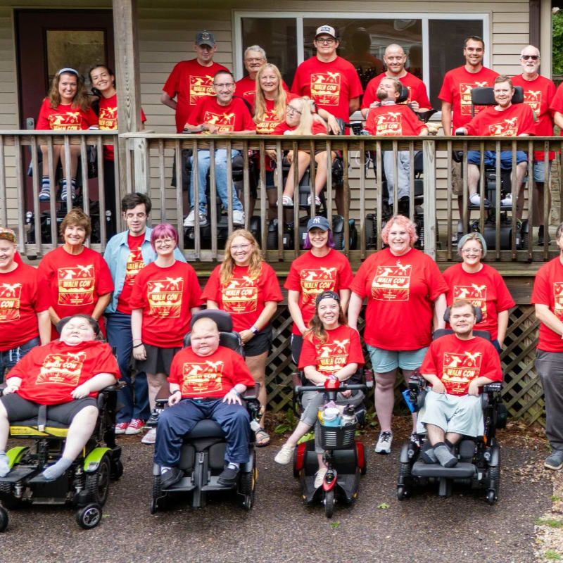 A group of people in matching red shirts pose for a photo on a porch, with several individuals in wheelchairs in the foreground.