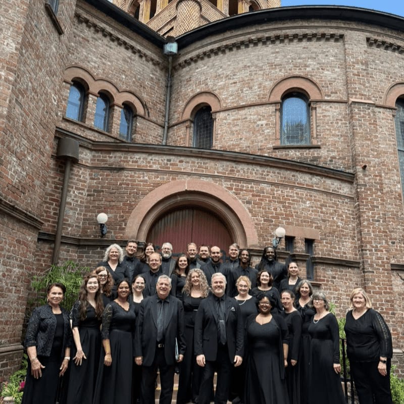 A choir in black attire stands on steps in front of a large brick building with arched windows.