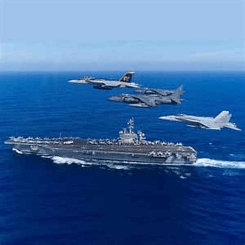 A large aircraft carrier sails through the ocean with three fighter jets flying overhead against a clear blue sky.