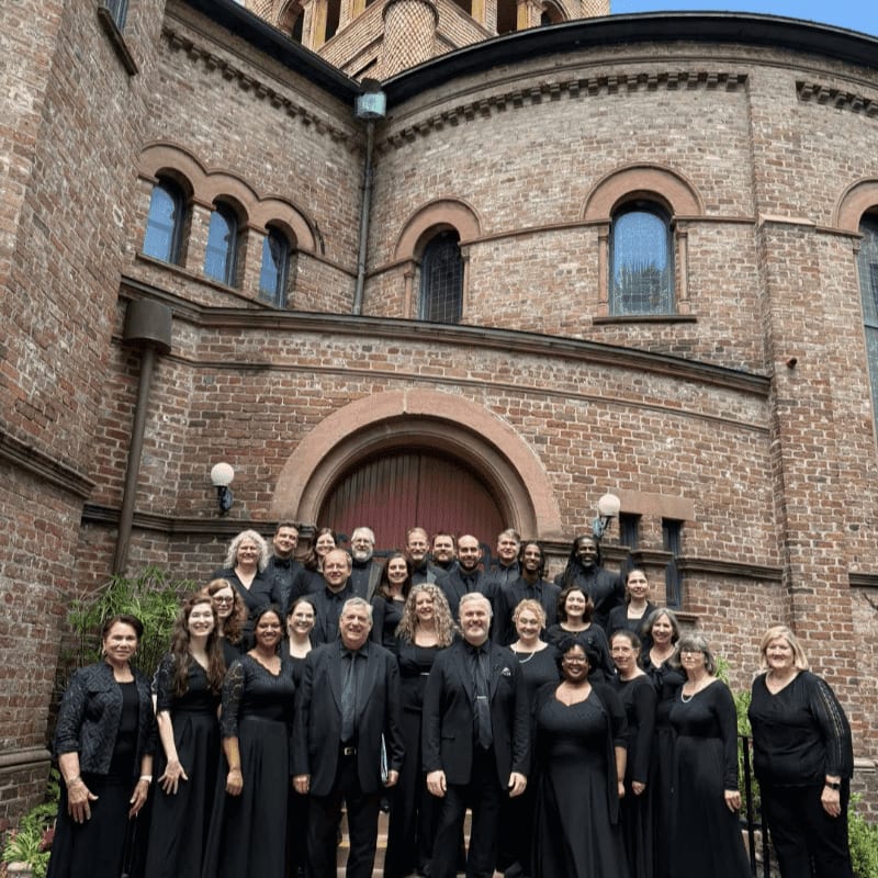A choir in black attire stands on steps in front of a large brick building with arched windows.