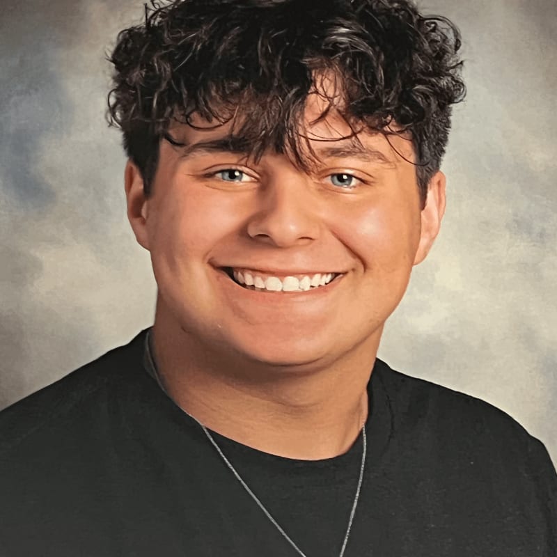 A smiling young man with curly dark hair and blue eyes in a black t-shirt is set against a mottled gray and blue background.