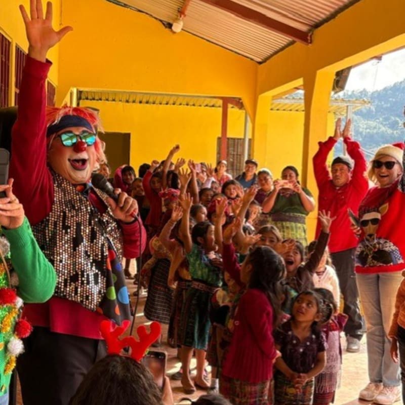 A clown with a microphone and a red bandana waves to a crowd of excited children with their hands raised in a yellow building.