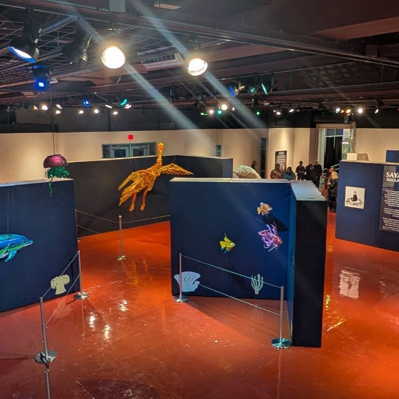 In the foreground, colorful marine life models are displayed on dark blue panels with a shiny red floor, while the background reveals a dimly lit exhibition hall with people observing the displays.
