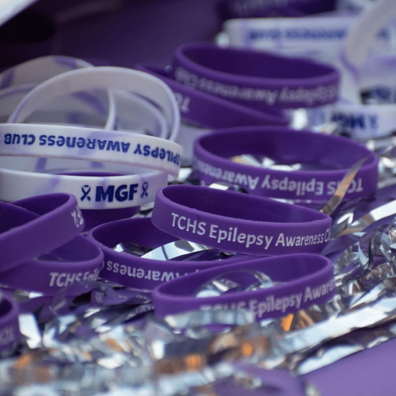 Purple and white "Epilepsy Awareness" wristbands are scattered on a shiny, crinkled surface in the foreground, with more wristbands visible in the blurred background.