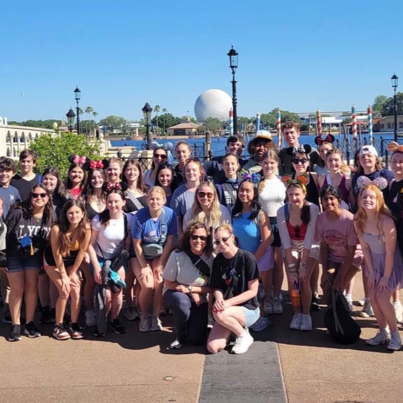 A group of young people pose for a photo in front of a large geodesic dome and a body of water, with a clear blue sky overhead.