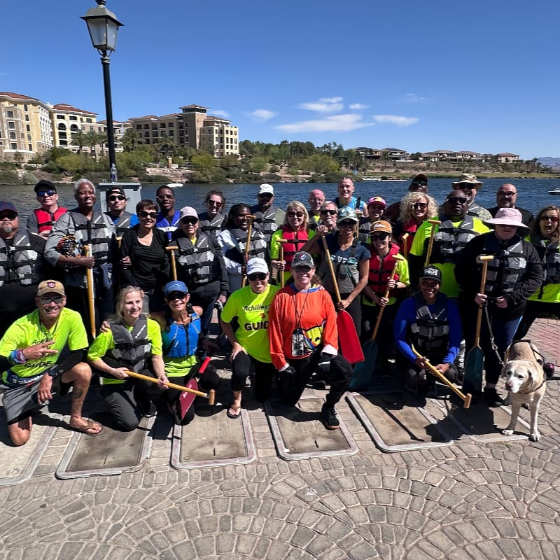 A group of people in life vests pose for a photo on a paved dock in front of a lake with buildings and hills in the background.